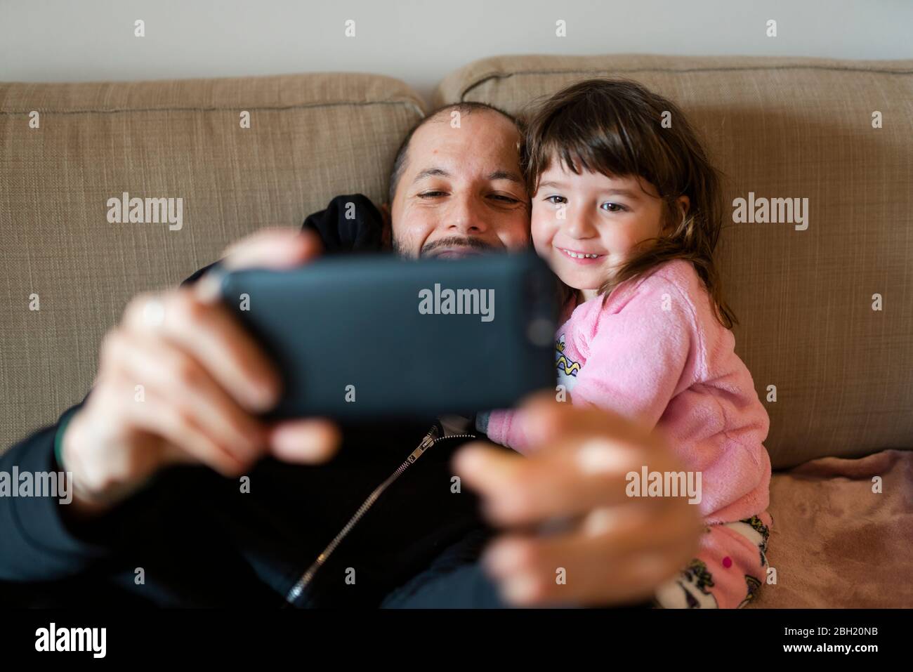 Feliz padre e hija haciendo una llamada de vídeo a la familia con el teléfono móvil, sentado en el sofá Foto de stock