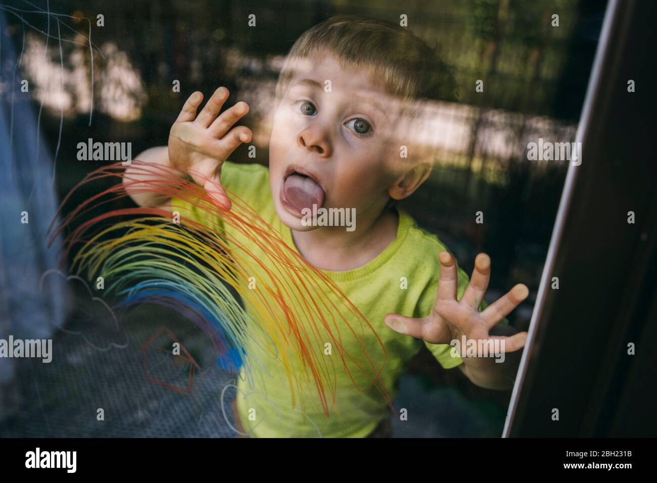Retrato de un niño parado detrás de la puerta del balcón que lamiendo el cristal con el arco iris dibujado Foto de stock