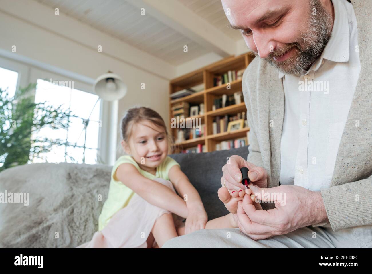 Padre pintando las uñas de sus hijas con esmalte de uñas rojo en el sofá de la sala de estar Foto de stock