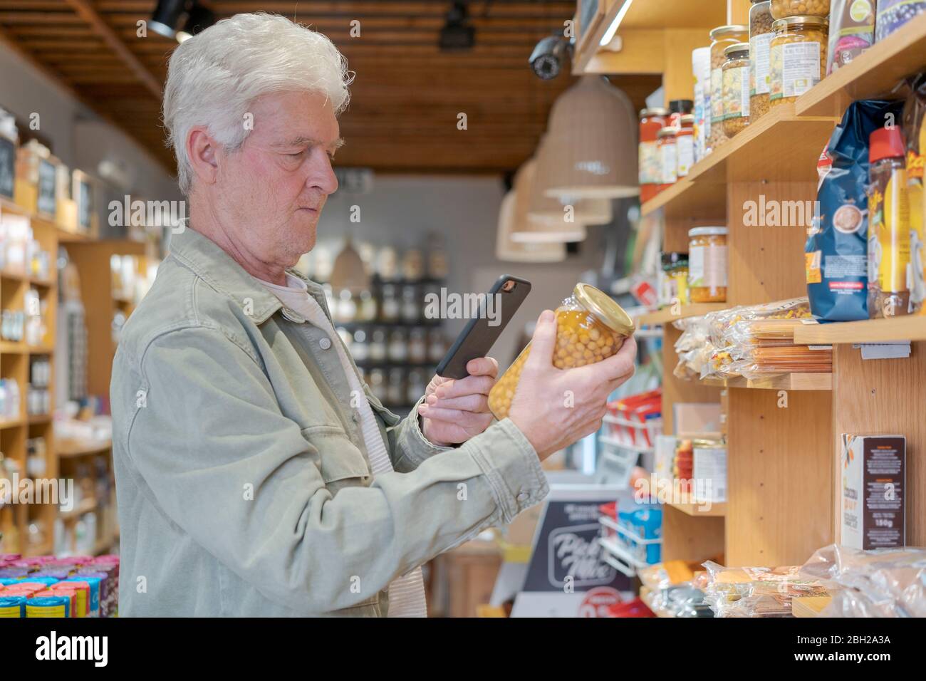 Hombre mayor con smartphone comprando comestibles en una pequeña tienda de alimentos Foto de stock