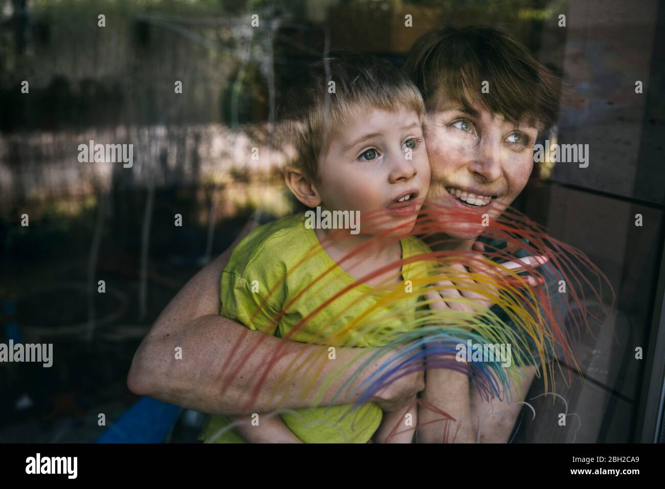 Retrato de madre feliz y su hijo pequeño mirando por la ventana con el arco iris dibujado Foto de stock