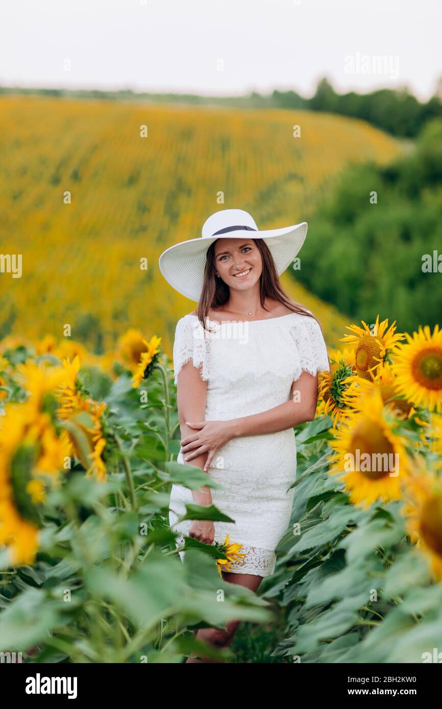 Feliz hermosa joven mujer en un vestido blanco y sombrero en un campo de  girasoles disfruta de la luz del sol. Paseo de verano en el campo de  girasoles de un joven