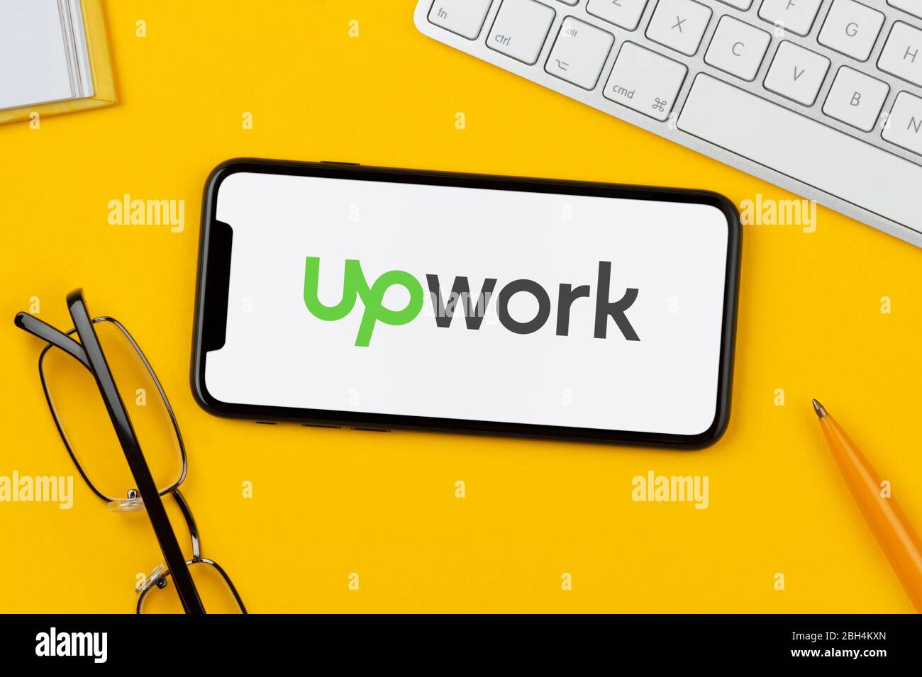 Un smartphone que muestra el logotipo de Upwork se apoya sobre un fondo amarillo junto con un teclado, gafas, lápiz y libro (sólo para uso editorial). Foto de stock