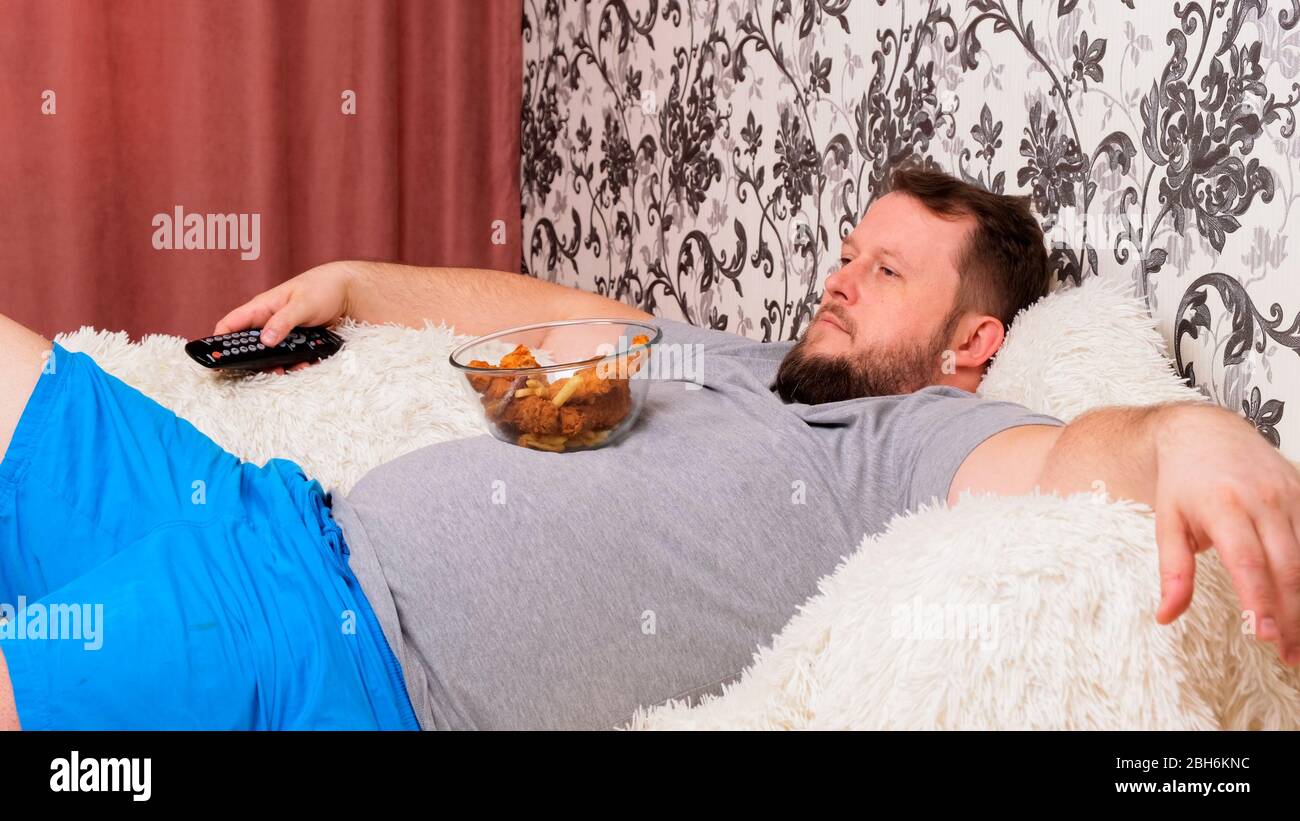 El hombre gordo con un vientre grande está acostado en el sofá con comida  rápida y se abate delante de la TV con el mando a distancia en su mano,  primer plano