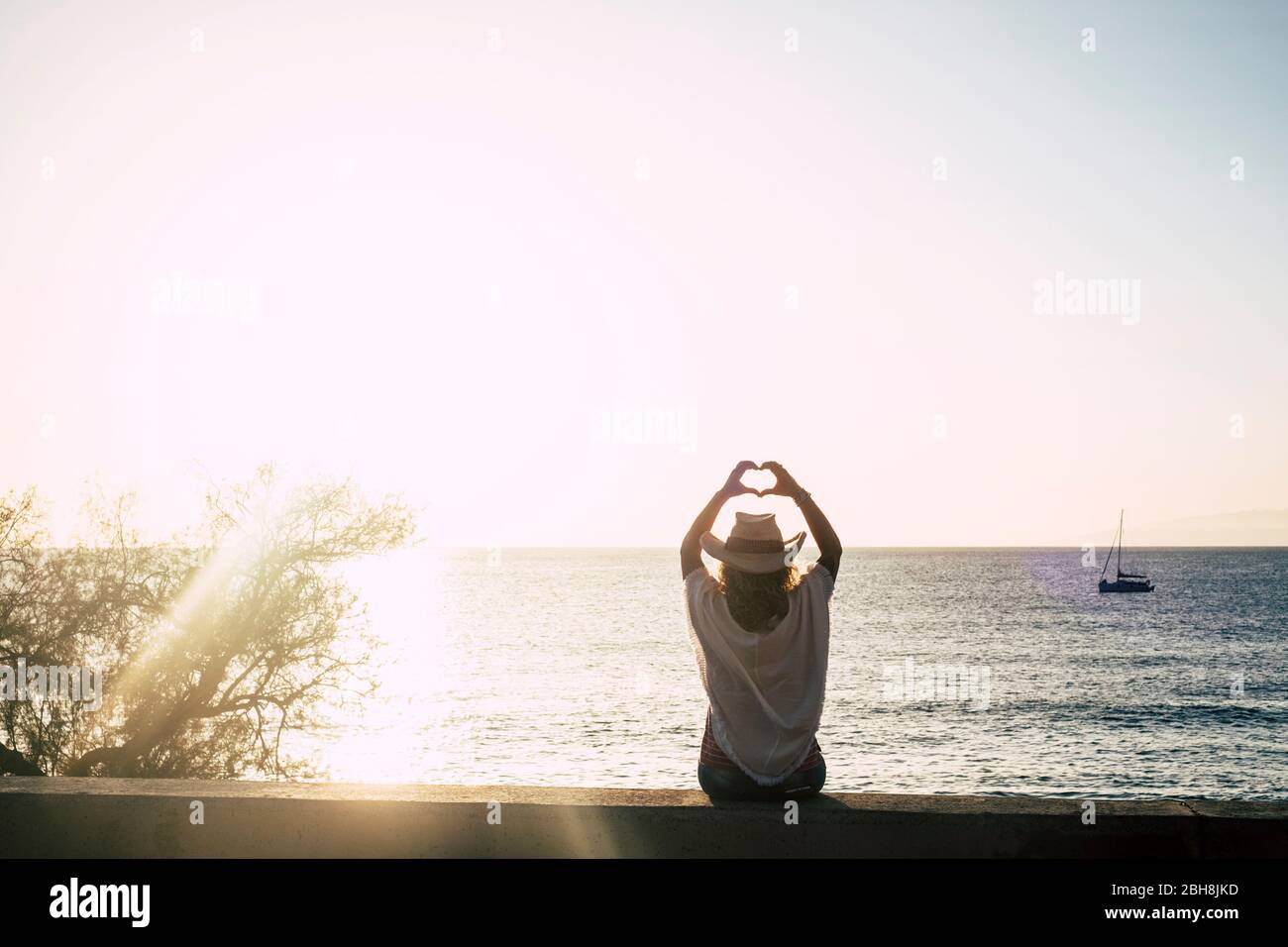 chica linda con un sombrero de estilo veraniego y cowbly visto desde atrás haciendo señas de hogar con las manos - luz de sol y océano con barco en el fondo - libertad y disfrute del concepto de estilo de vida - cielo despejado Foto de stock