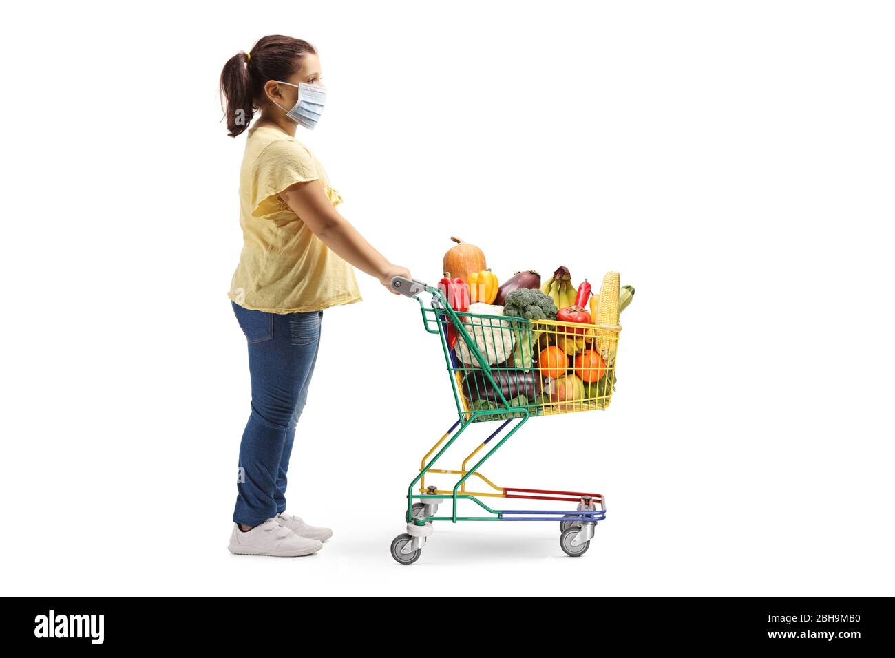 Foto de perfil completo de una chica de pie con un carrito de compras y con una máscara médica aislada sobre fondo blanco Foto de stock