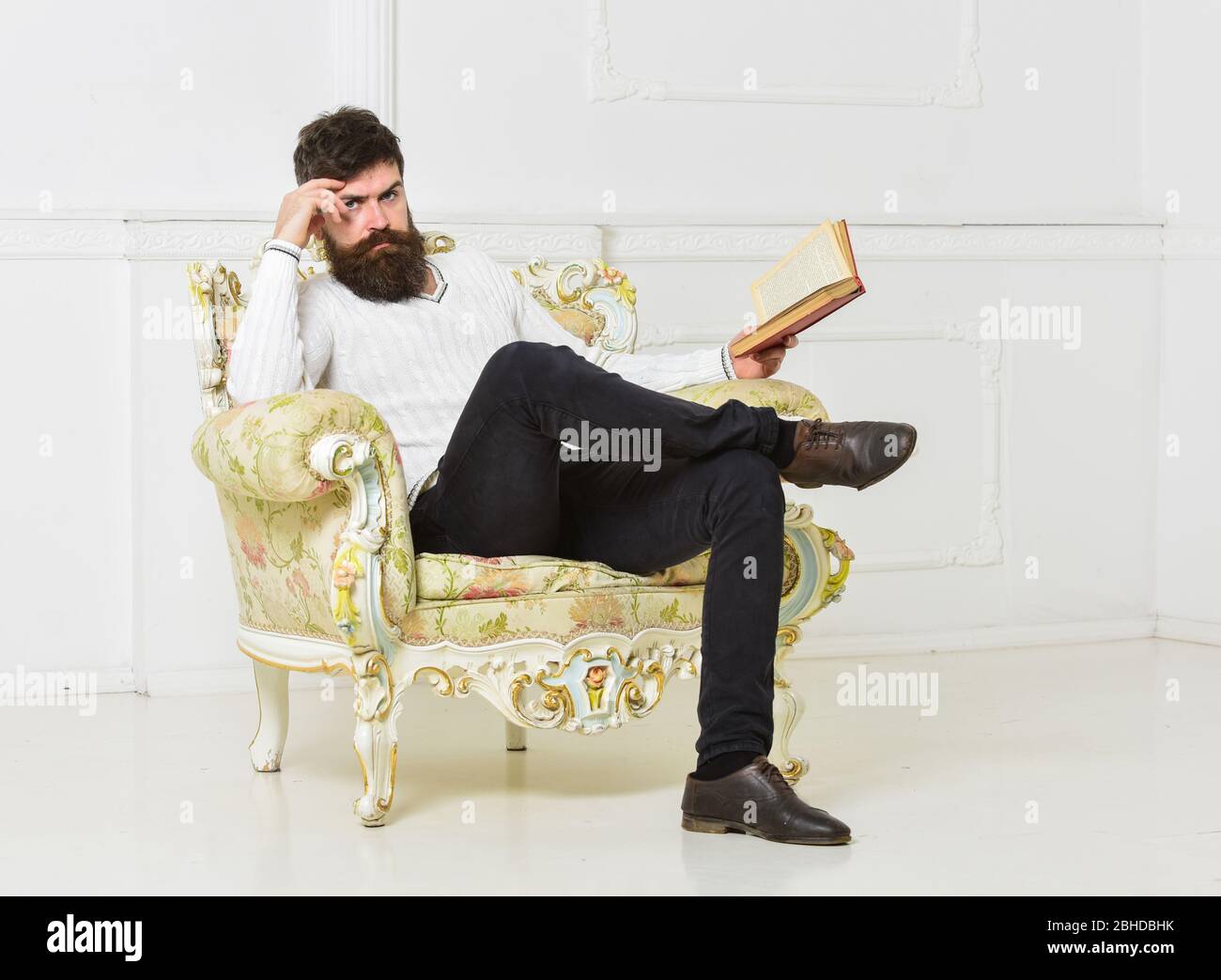 Hombre con barba y bigote sentado en el sillón, sostiene el libro, fondo  blanco de la pared. Reflexiones sobre el concepto de literatura. Conocedor  de cara pensativa terminó leyendo el libro. Tipo