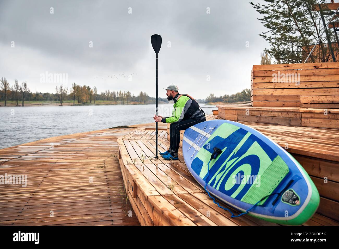 Atleta de neopreno con paddle surf y sentado en el muelle de madera cerca del lago Foto de stock