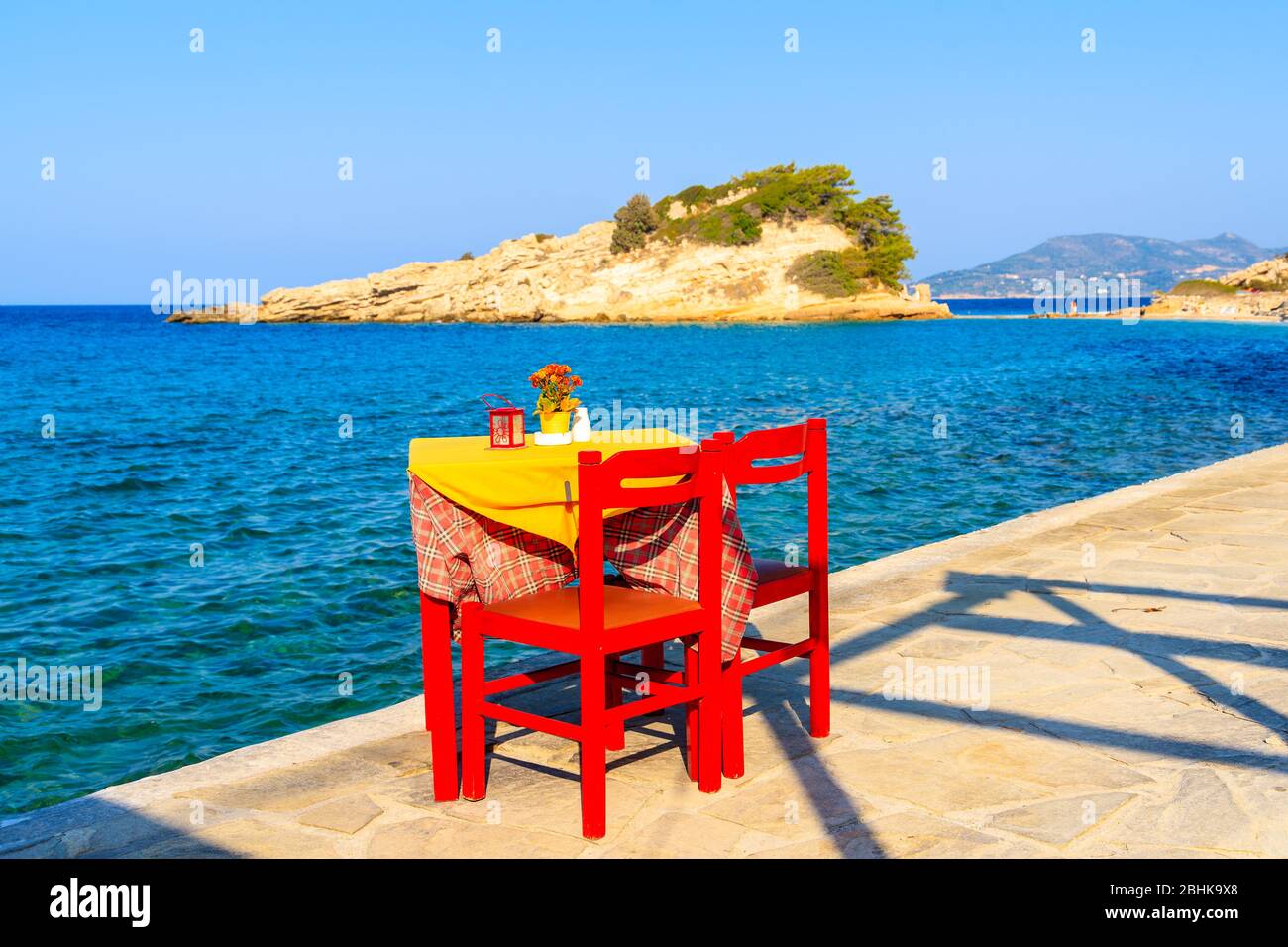 Mesa Taverna con sillas rojas en el paseo marítimo costero con el mar en el fondo en el pueblo de Kokkari, isla de Samos, Grecia Foto de stock