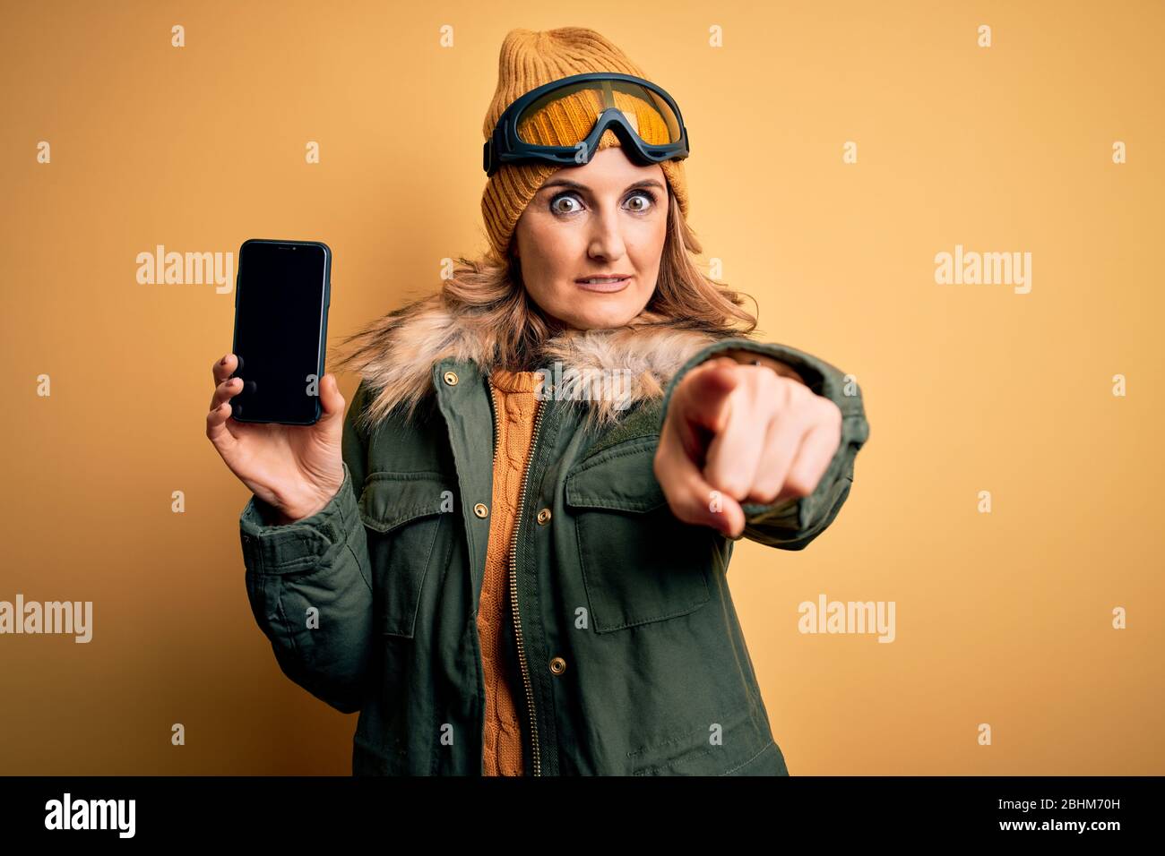 Mujer rubia de edad media con gafas de esquí sosteniendo un smartphone mostrando la pantalla apuntando con el dedo a la cámara y a usted, signo de mano, positivo Foto de stock