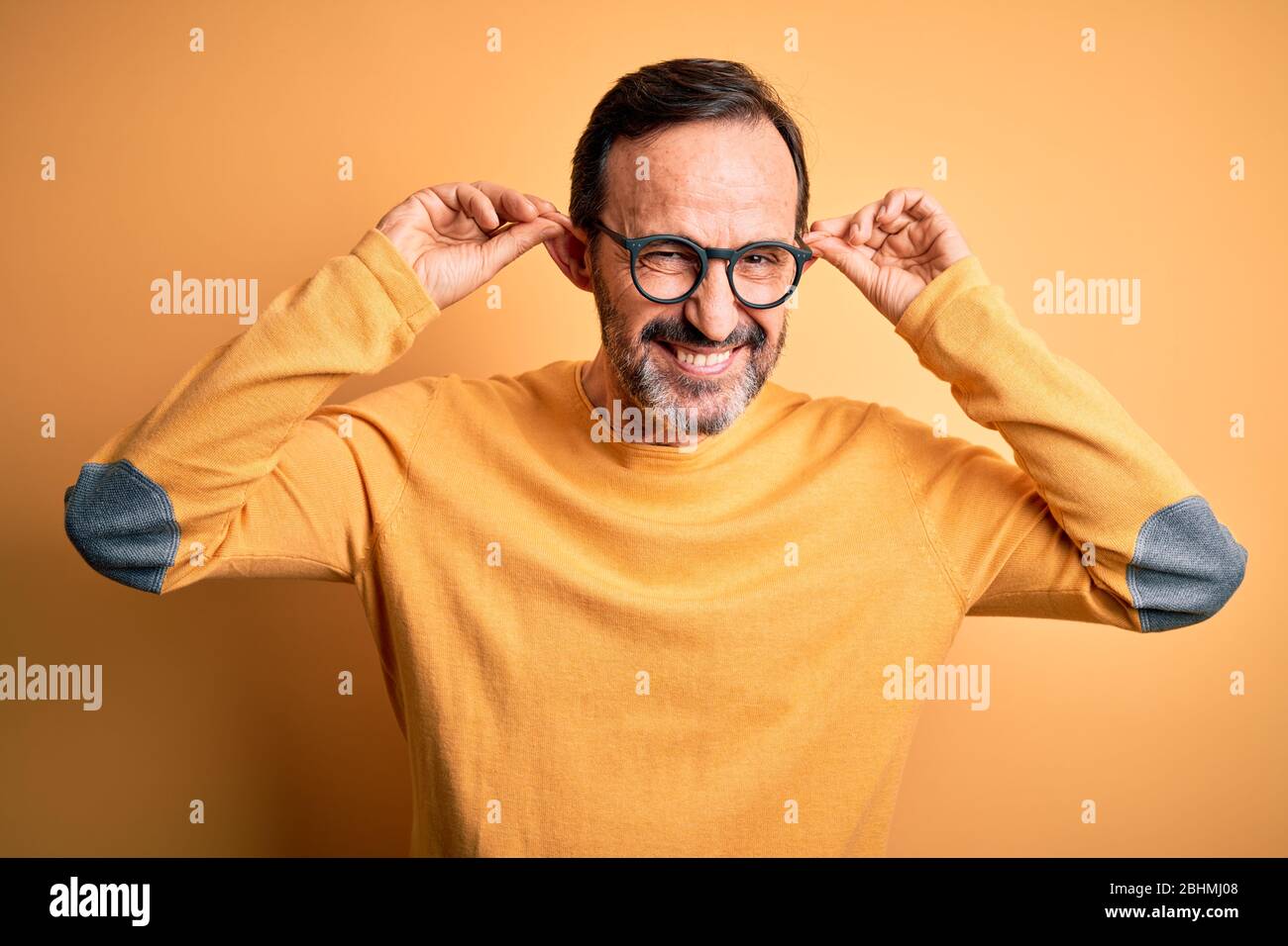 Edad media hombre caloroso con suéter informal y sobre fondo amarillo aislado Esmordete las orejas tirando con los dedos, gesto divertido. Audición de stock - Alamy