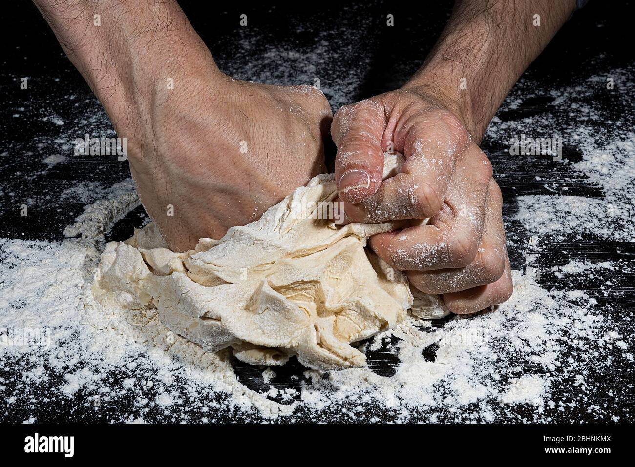 Puño de un hombre presionando una bola de harina amasada para hacer pan sobre una superficie negra llena de harina Foto de stock