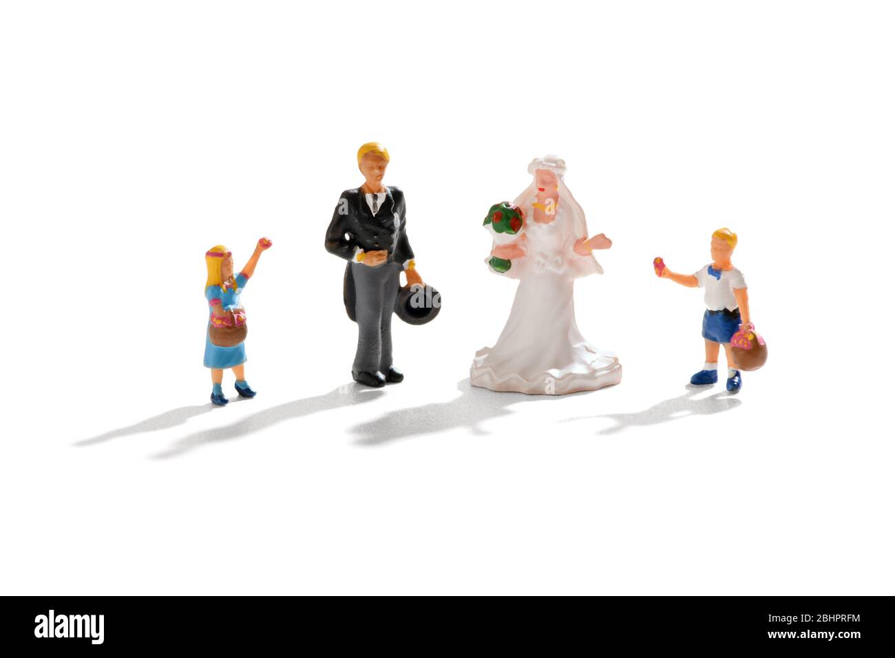 Mini pareja recién casados con la novia y novio de pie entre dos niños pequeños, una niña de flores y el niño de la página en una escena de boda sobre blanco con Foto de stock