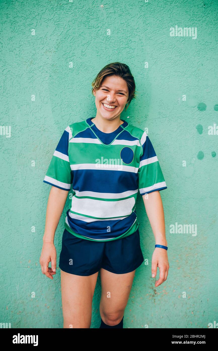 Una mujer que lleva una camiseta de rugby azul, blanca y verde, que está de pie contra una pared verde mirando a la cámara y sonriendo Fotografía de stock -