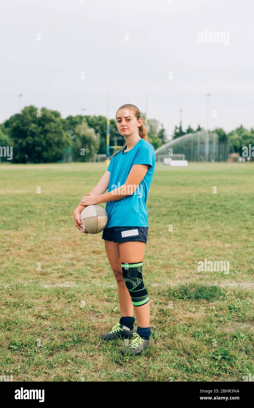 Una mujer de pie un campo de entrenamiento con pantalones cortos y camiseta con una correa para la rodilla que sostiene un balón de rugby Fotografía stock -