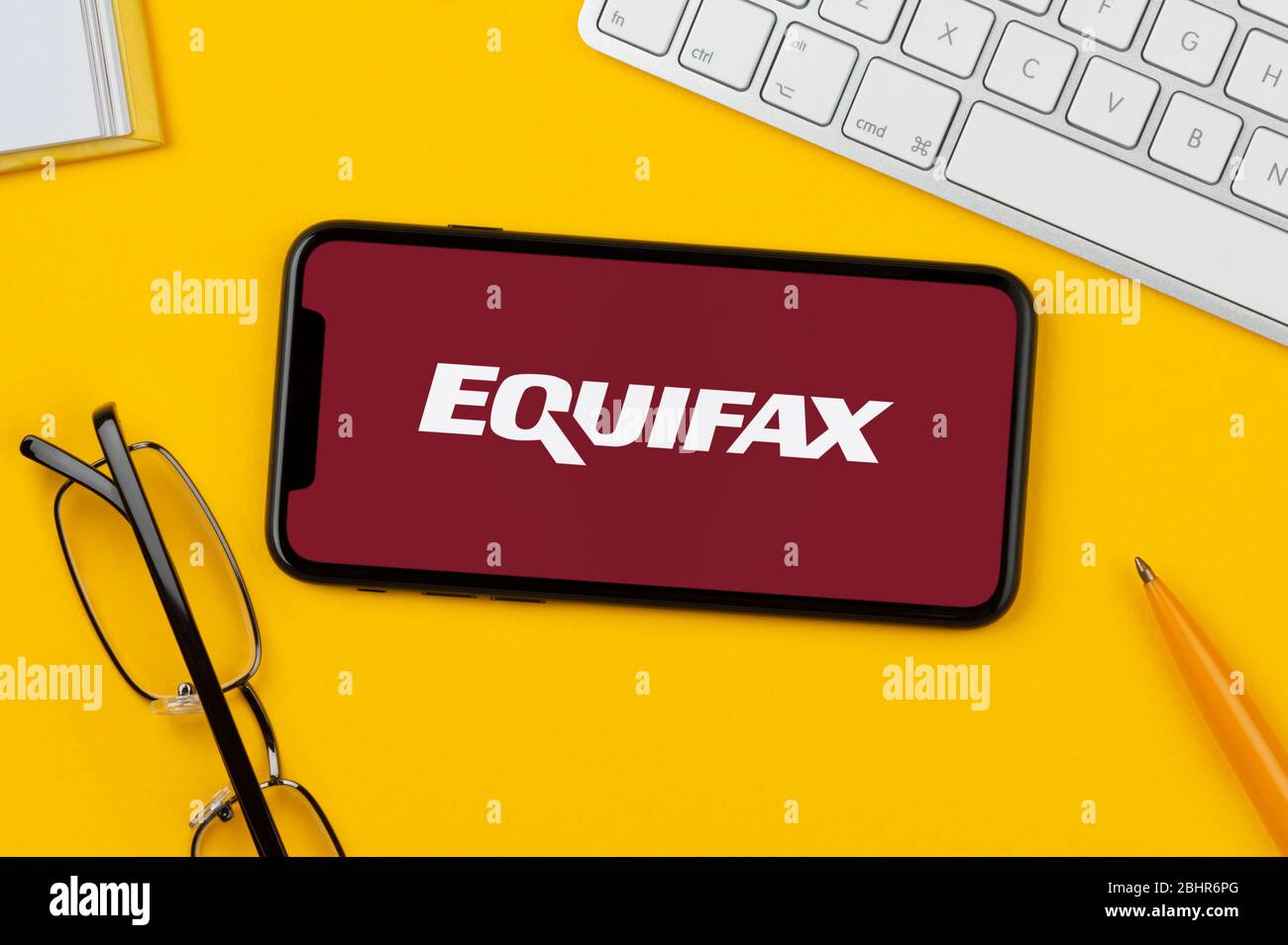 Un smartphone que muestra el logotipo de Equifax descansa sobre un fondo amarillo junto con un teclado, gafas, lápiz y libro (sólo para uso editorial). Foto de stock
