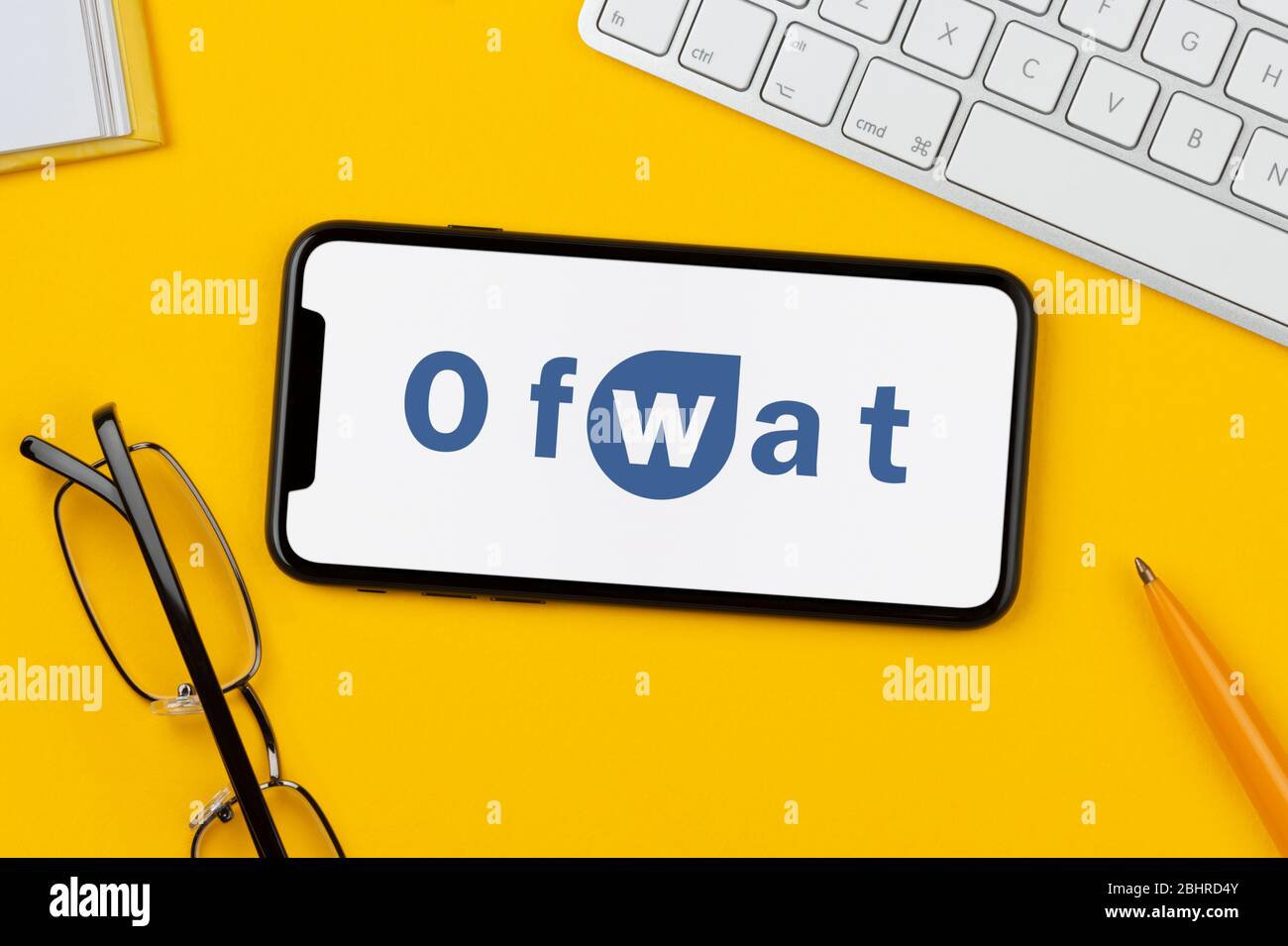 Un smartphone que muestra el logotipo de Ofwat se apoya sobre un fondo amarillo junto con un teclado, gafas, lápiz y libro (sólo para uso editorial). Foto de stock