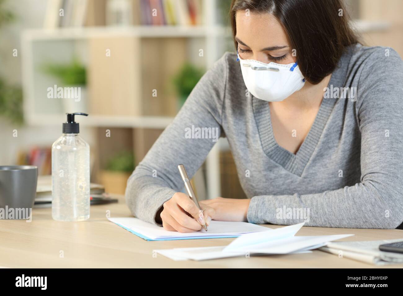Mujer con máscara protectora debido coronavirus escribiendo carta sentada en un escritorio en casa Foto de stock