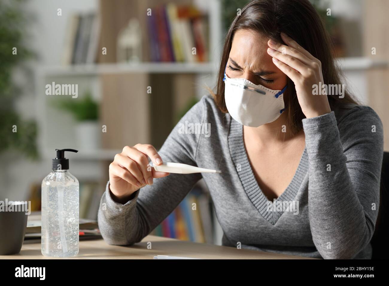 Mujer enferma con máscara protectora que sufre de síntomas de coronavirus con dolor de cabeza sosteniendo el termómetro por la noche en casa Foto de stock