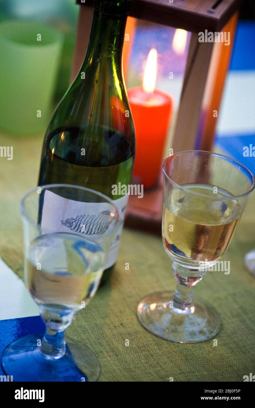 Mesa puesta para una fiesta en el exterior en una noche de verano, vasos de vino blanco con media botella vacía y vela roja en farol de madera. - Foto de stock