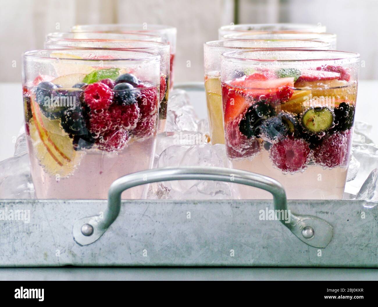 Agua con gas con fruta fresca y bayas en vasos sentados sobre hielo picado en la bandeja de metal. Foto de stock