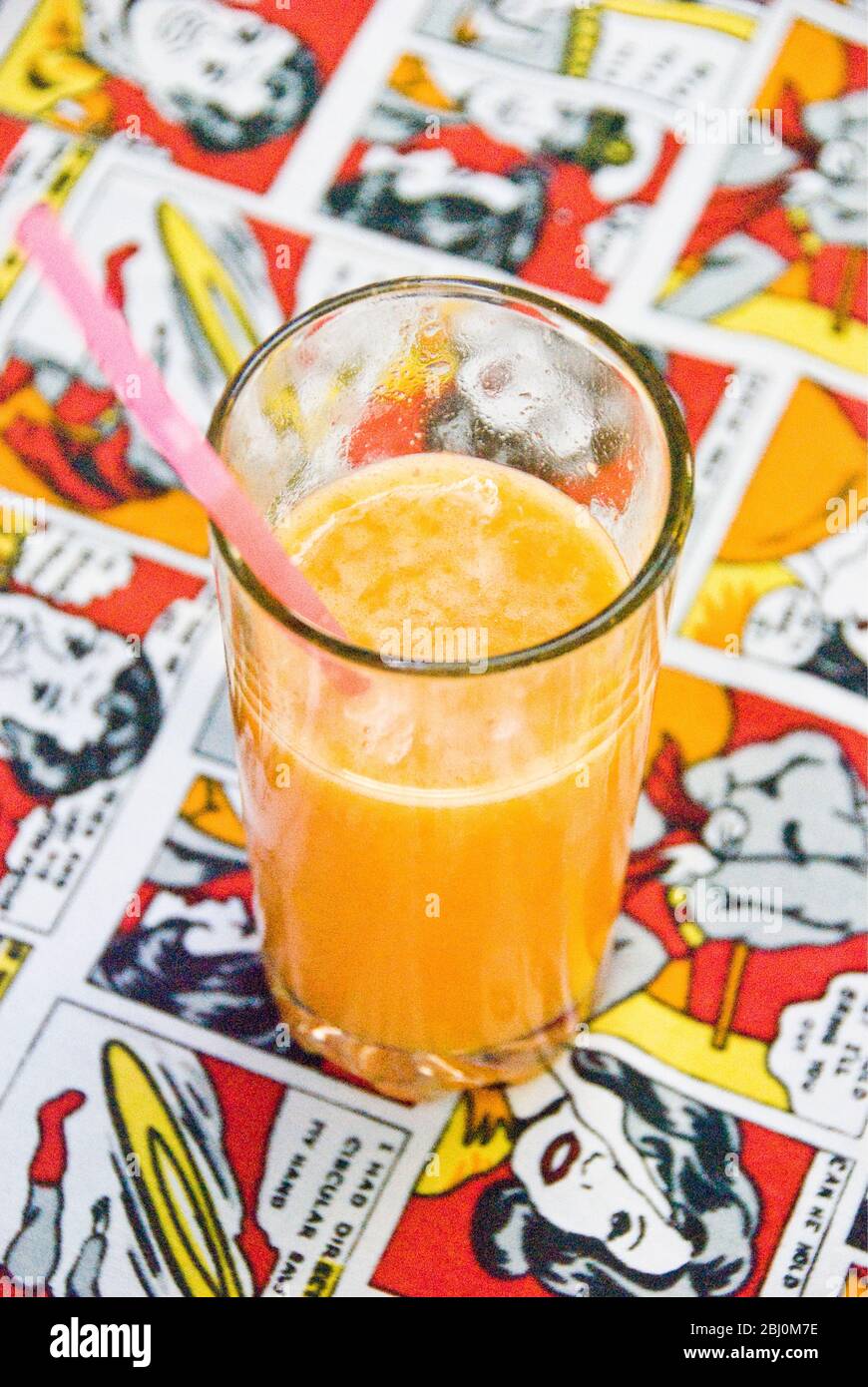 Zumo de naranja recién exprimido en un café de cristal sobre tela de mesa con un dibujo de tiras cómicas - Foto de stock