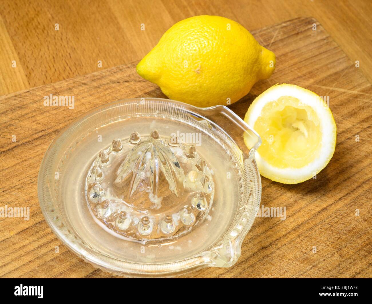 Zumo de limón de medio limón exprimido en un exprimidor de limón de cristal con un limón entero sobre una tabla de madera Foto de stock
