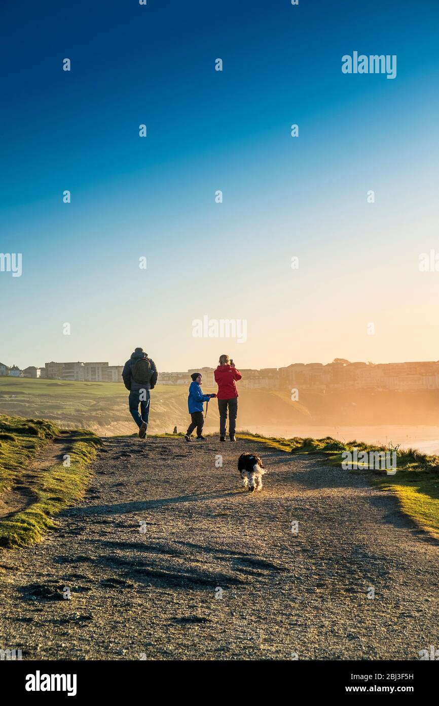 Una familia y su perro mascota caminando por el sendero costero mientras el sol se pone sobre la Bahía Fistral en Newquay en Cornwall. Foto de stock