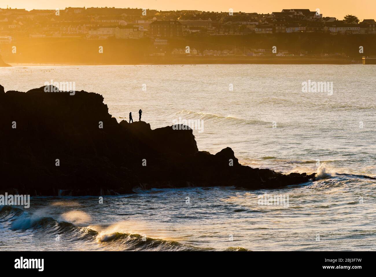 Dos figuras en las rocas siluetas por el sol que pone en Newquay en Cornwall. Foto de stock