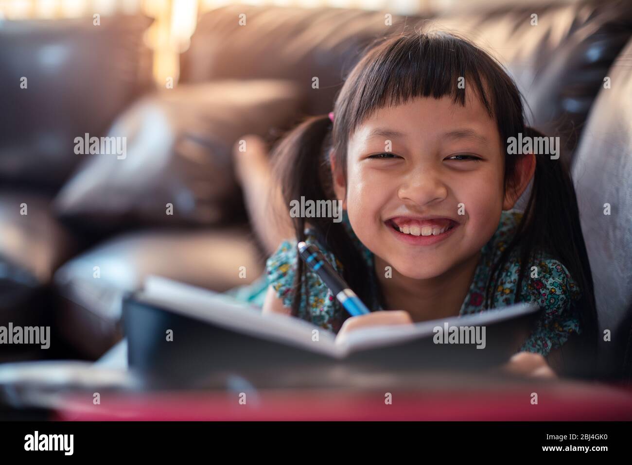 Niña pequeña y feliz acostada en el sofá con aprender por ordenador portátil y escribir un libro en casa, distancia social durante la cuarentena, concepto de educación en línea Foto de stock