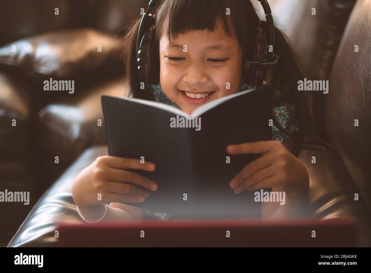 Niña pequeña y feliz acostada en el sofá con aprender por ordenador portátil y escribir un libro en casa, distancia social durante la cuarentena, concepto de educación en línea Foto de stock