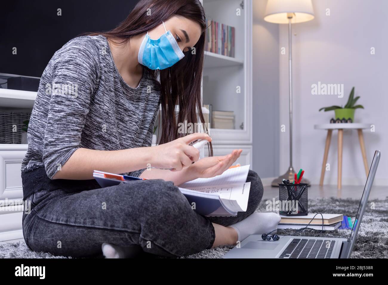 Niña de la escuela secundaria con máscara en la cara usando desinfectante antes de hacer su tarea. Educación en línea durante la pandemia del coronavirus. Foto de stock