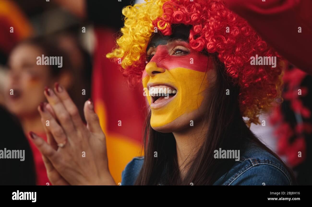 Mujer sentada en el estadio con una peluca y su cara pintada con colores de bandera alemana aplaudiendo a su equipo. Mujer de Alemania en zona de fans viendo un so Foto de stock