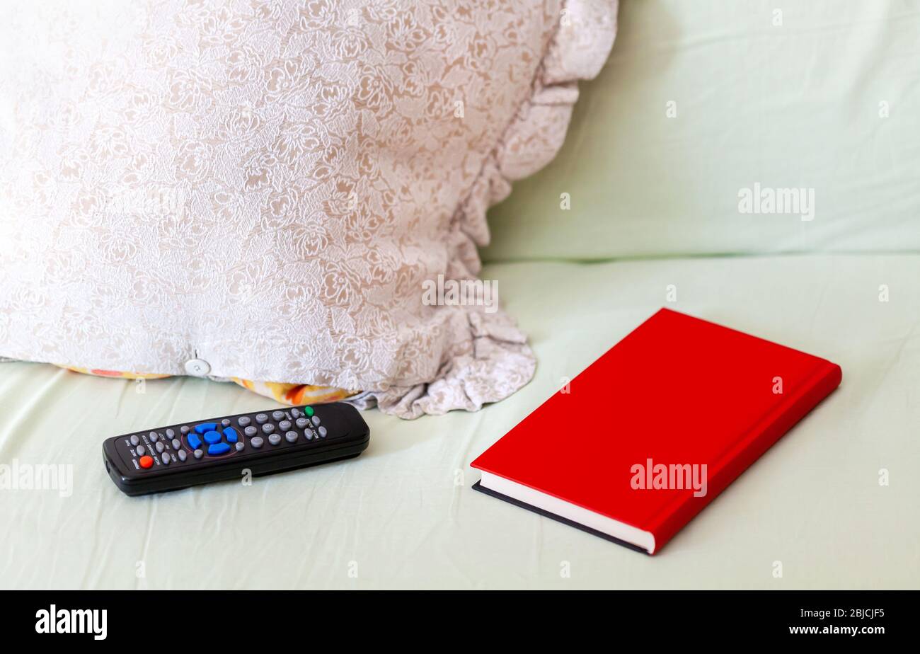 Mando a distancia para TV, libro rojo y un juego de almohadas de objetos sobre una cama no hecha / sofá Ocio, concepto de tiempo libre, actividades interiores, aburrimiento Foto de stock