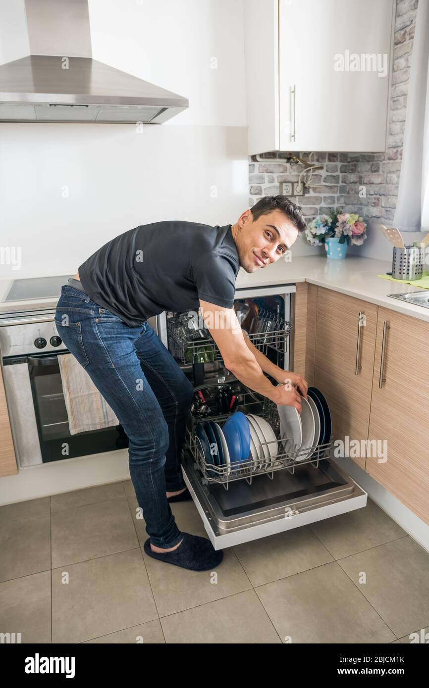 Hombre sacando platos del lavavajillas en la cocina y mirando la cámara  sonriendo. Cuerpo completo Fotografía de stock - Alamy
