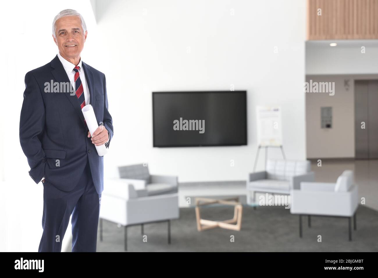 Hombre de negocios maduro sonriente, parado en un moderno vestíbulo del edificio de oficinas con monitor de pantalla plana. Él está sosteniendo un documento enrollado en su mano. Horizontal Foto de stock