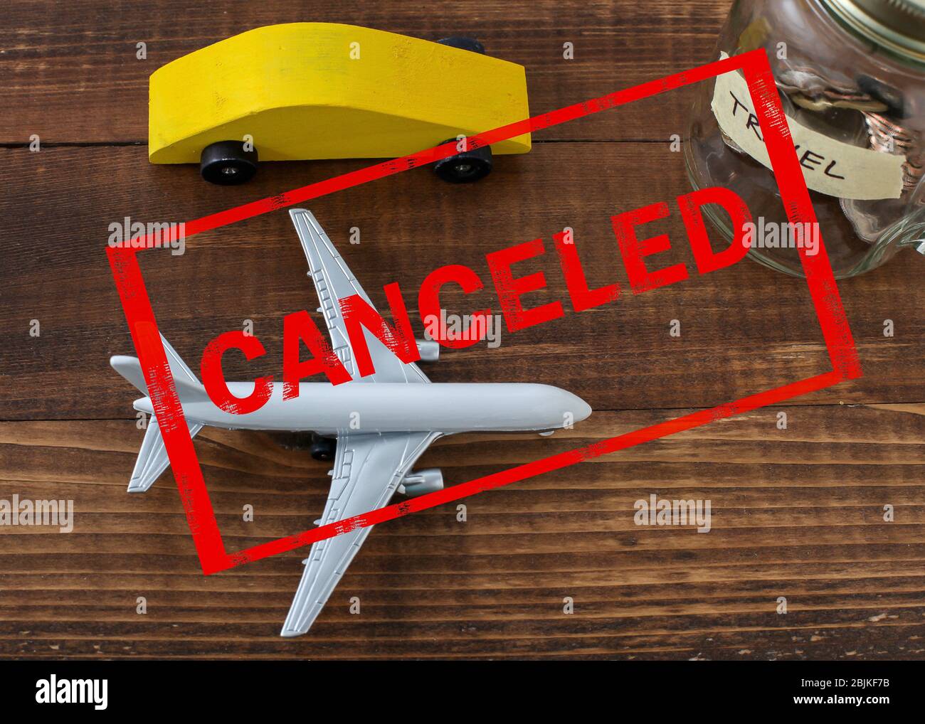 Coche de madera con avión y monedas en tarro con etiqueta de viaje y texto rojo cancelado sobre la imagen. Foto de stock