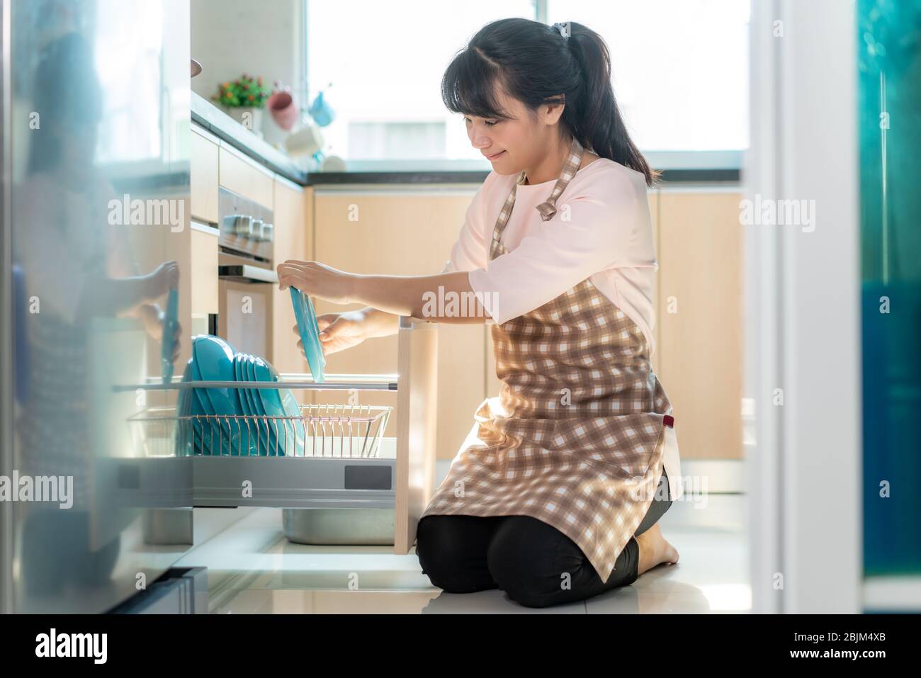 Atractiva joven asiática cargando el lavavajillas en armarios de la cocina mientras se hace la limpieza en casa durante su estancia en casa utilizando el tiempo libre abou Foto de stock