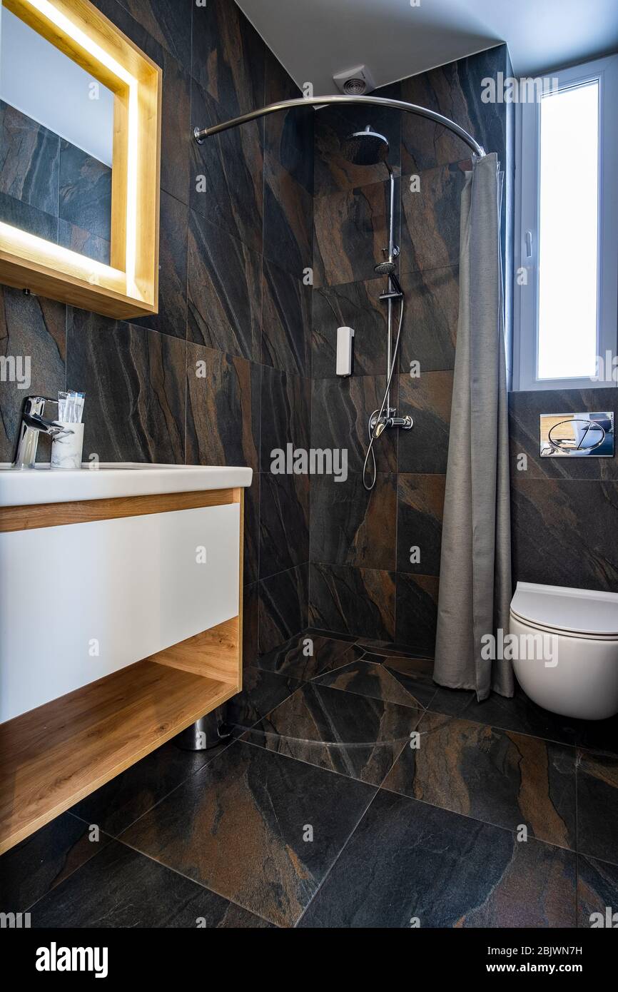 Interior de un moderno y elegante cuarto de baño con paredes de azulejos  negros, ducha de cortina y muebles de madera con lavabo y un gran espejo  iluminado Fotografía de stock -