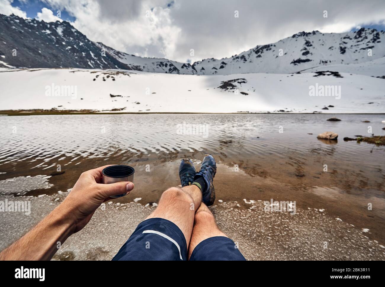 Taza de Té y las piernas del hombre en zapatos de Seguimiento y vista sobre el lago glaciar cubierto de nieve en las montañas Foto de stock