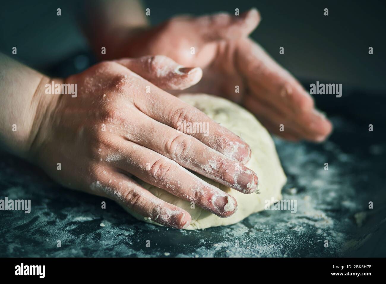 El cocinero amasa masa de pan con las manos fuertes en una bandeja oscura llena de harina desmenuzada blanca, iluminada por la luz. Foto de stock
