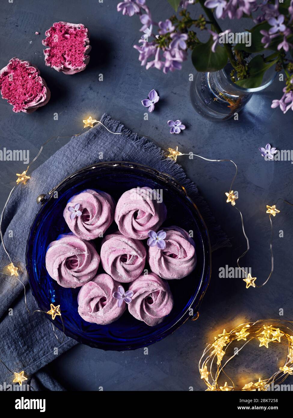 Zephyr o Marshmallow caseros dulces violeta atmosférico de grosella negra cerca de flores lilas y estrellas mágicas luces en la vista superior de fondo oscuro Foto de stock