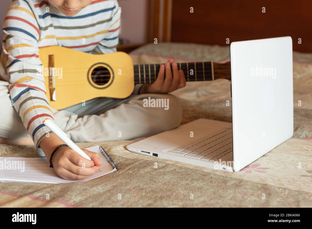 El niño trata de aprender la lección de la timple en casa del Internet con  su computadora portátil. Escribe una nueva canción con acordes en su  cuaderno. El timple es un Fotografía