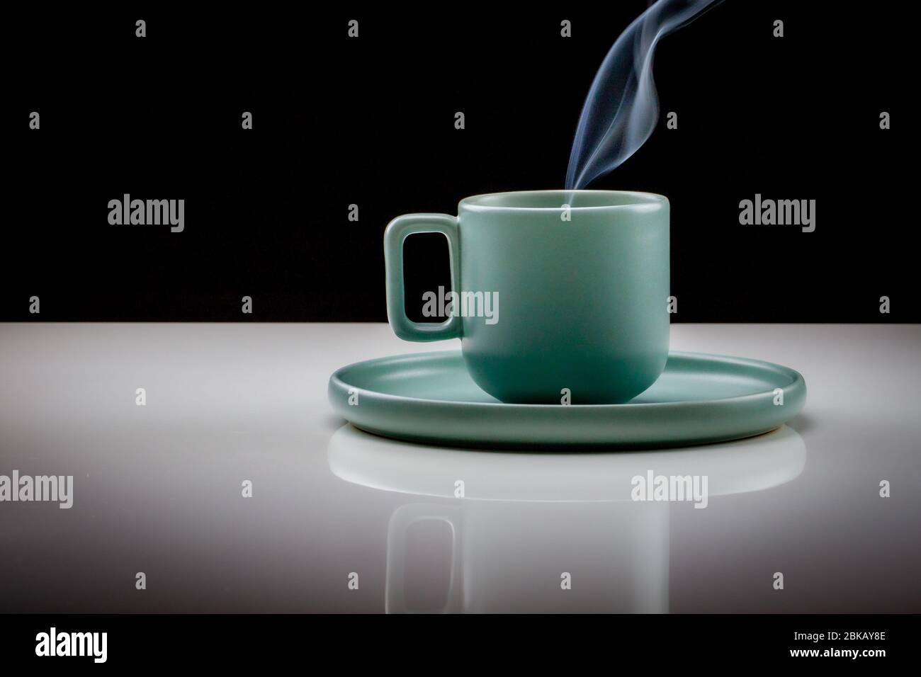 Taza de café o té de color verde pastel con líquido caliente, humo y vapor, fondo negro. Copa de pie sobre una mesa ligeramente reflectante que se desvanece Foto de stock
