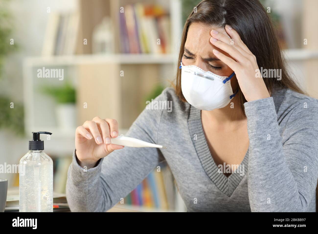 Mujer enferma con covid-19 síntomas que sufren dolor de cabeza sosteniendo el termómetro usando la máscara en casa Foto de stock