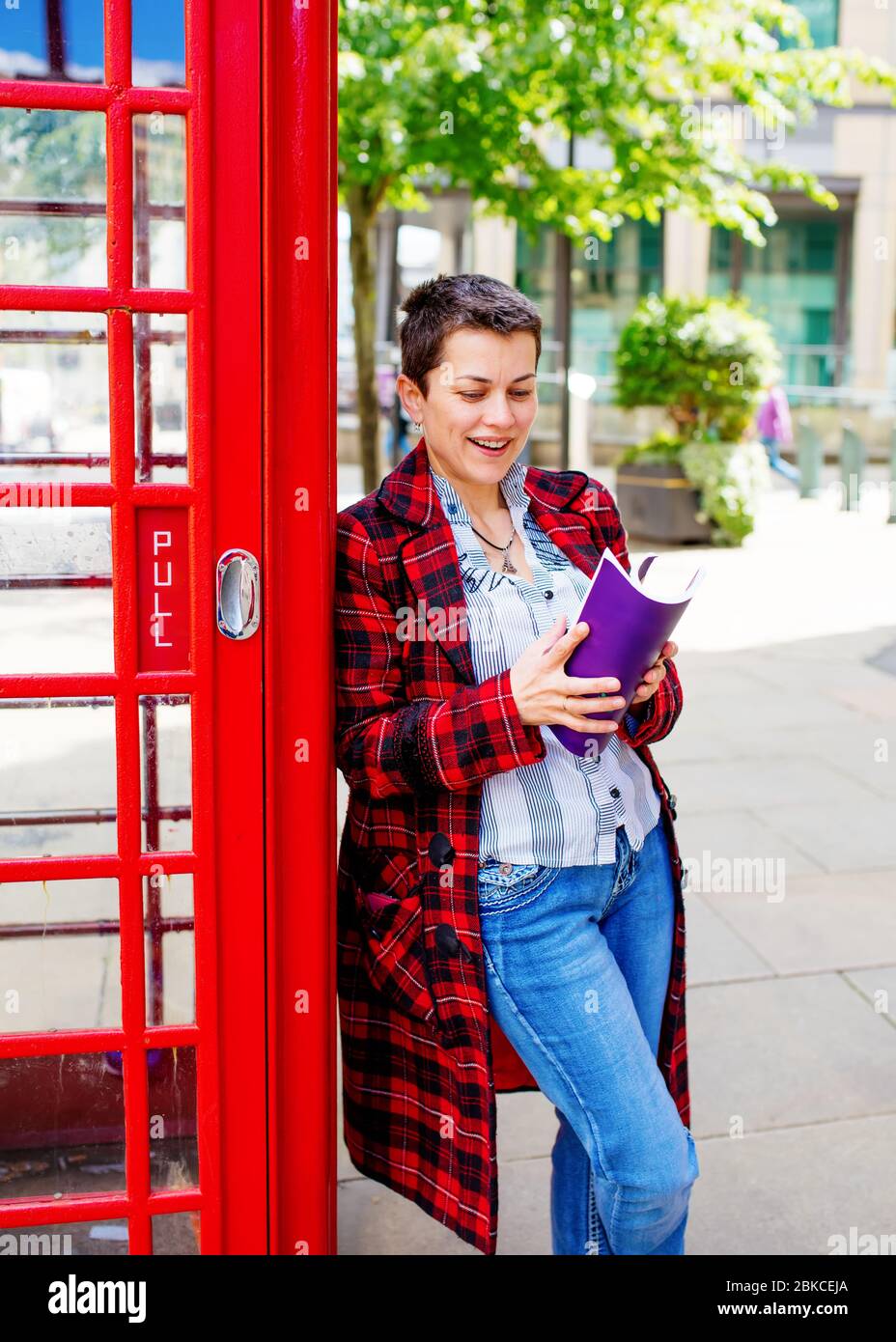 mujer con un abrigo rojo, jeans y camisa blanca sosteniendo el libro se  inclinó contra la caja roja del teléfono Fotografía de stock - Alamy