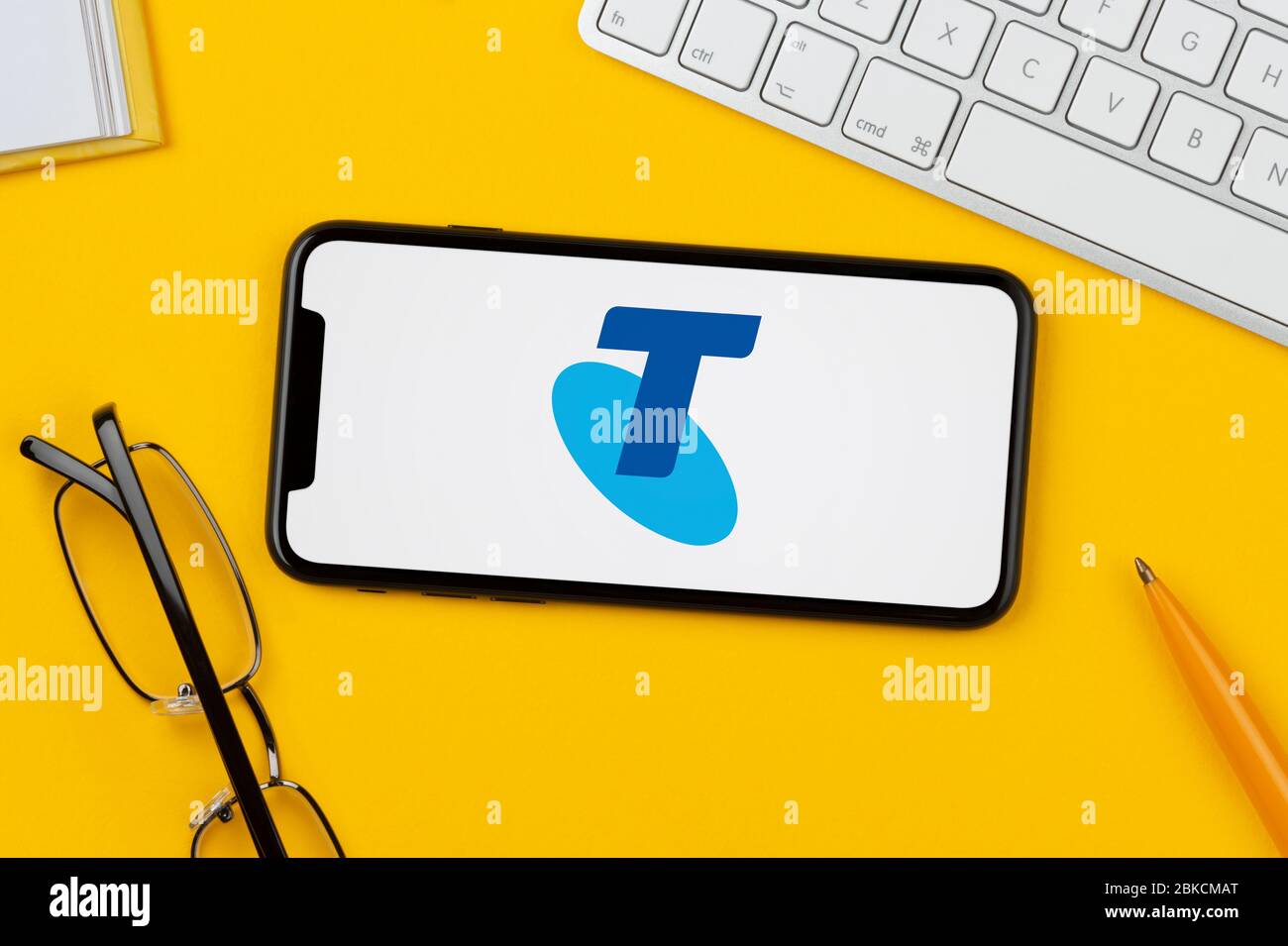 Un smartphone que muestra el logotipo de Telstra descansa sobre un fondo amarillo junto con un teclado, gafas, lápiz y libro (sólo para uso editorial). Foto de stock