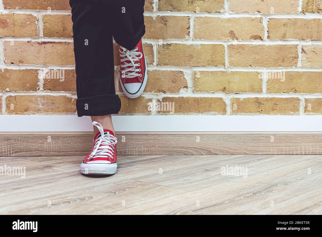 Mujer piernas en jeans y zapatillas de deporte rojas, mujer de pie frente la pared de ladrillo, concepto de Fotografía de -