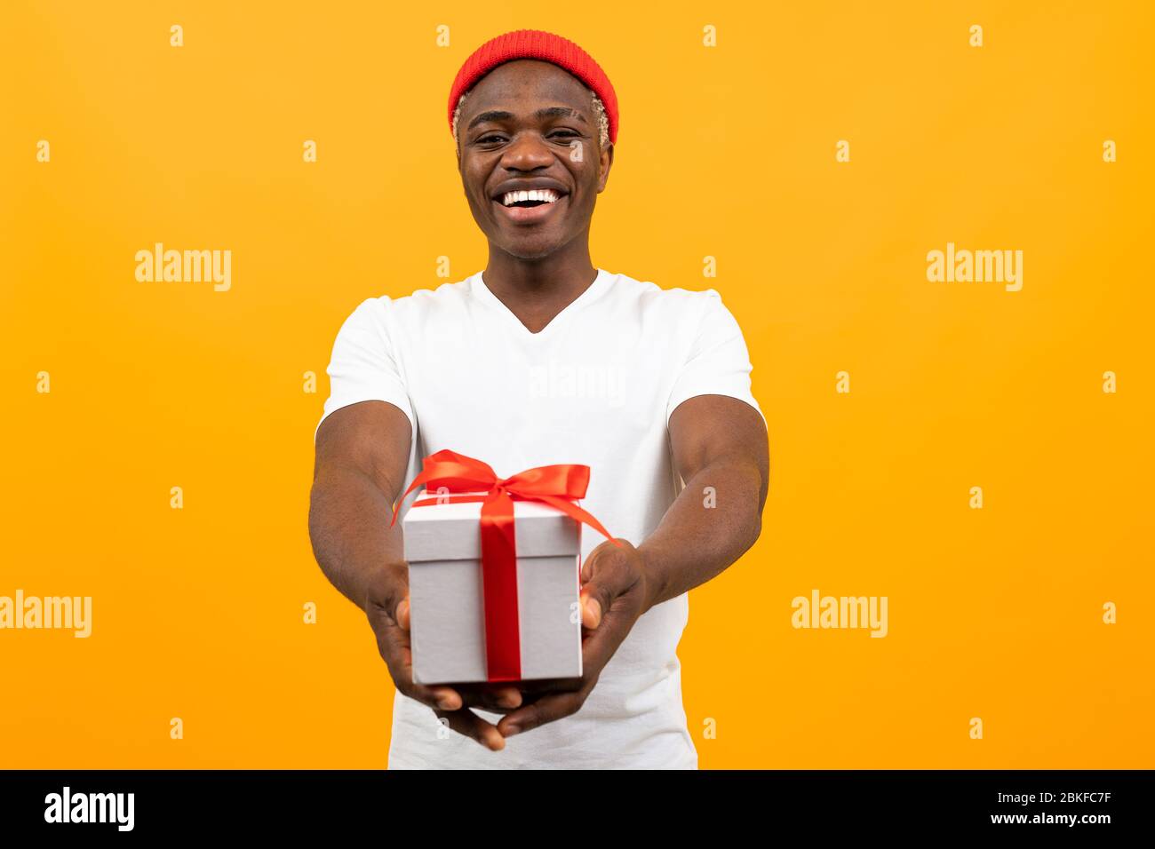 un hombre africano negro y coqueto con una sonrisa en una camiseta blanca  sostiene una caja de regalo con una cinta roja sobre un fondo amarillo con  espacio para copias Fotografía de