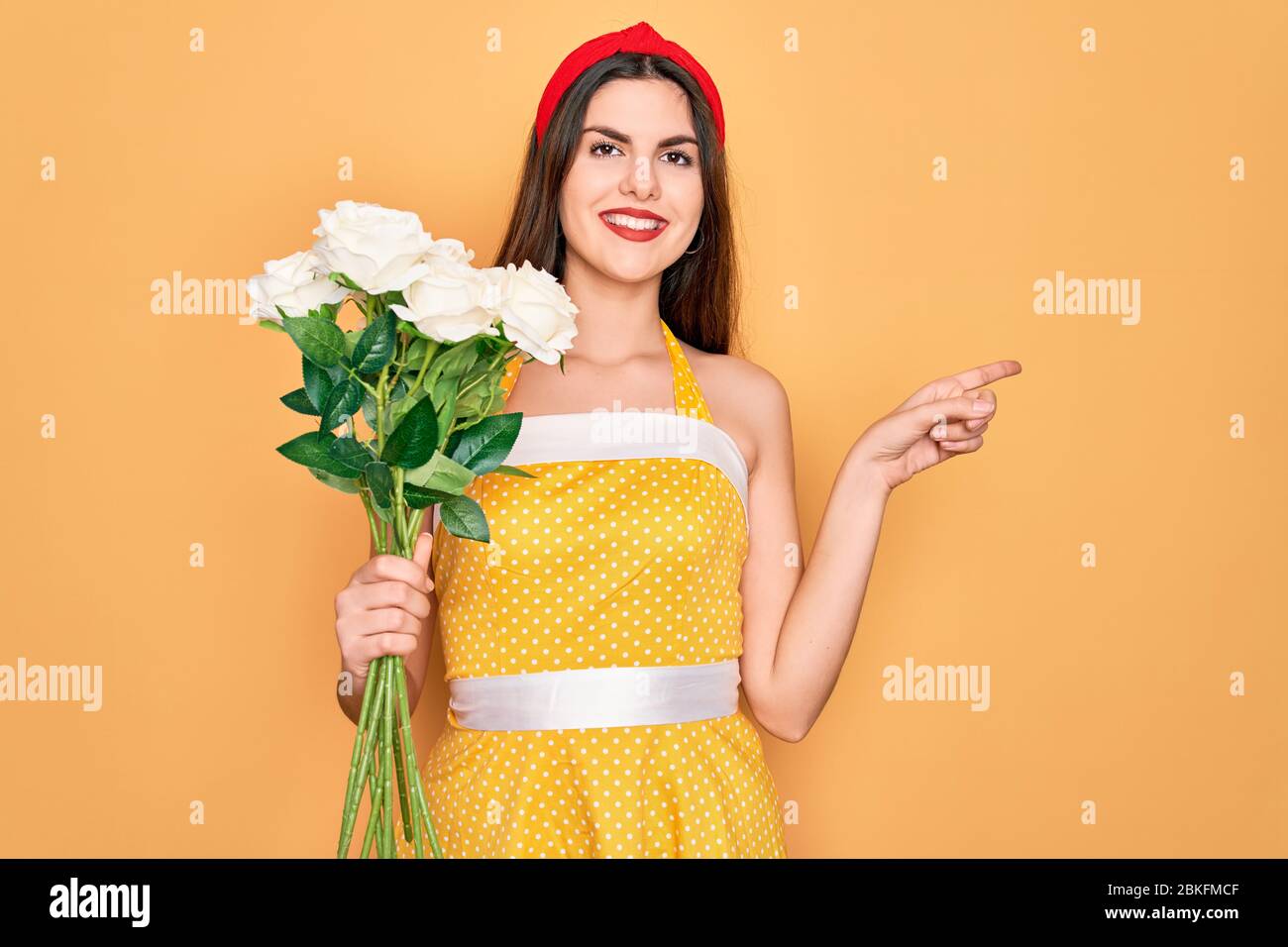 Joven hermoso pin up mujer con años 50 vestido de moda vintage celebración  de ramo de rosas flores muy feliz señalando con la mano y el dedo a un lado  Fotografía de